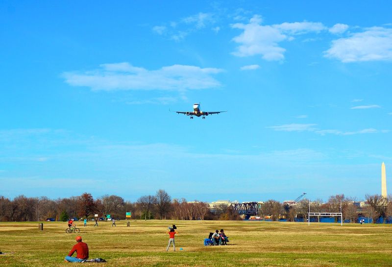 An image of the Gravelly Point Park on a sunny day when people are sitting on green grass and an aeroplane is flying from above